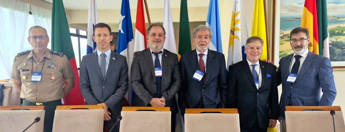 Inspetor-Geral com 5 pessoas atrás de uma mesa de conferência à frente de várias bandeiras nacionais visíveis ao fundo