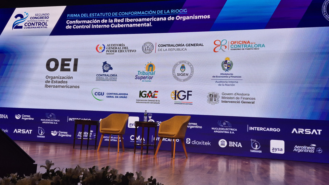 Palco do congresso onde se podem ver 2 cadeiras vazias e 2 mesas de apoio à frente de uma imagem gigante projetada do cartaz do congresso