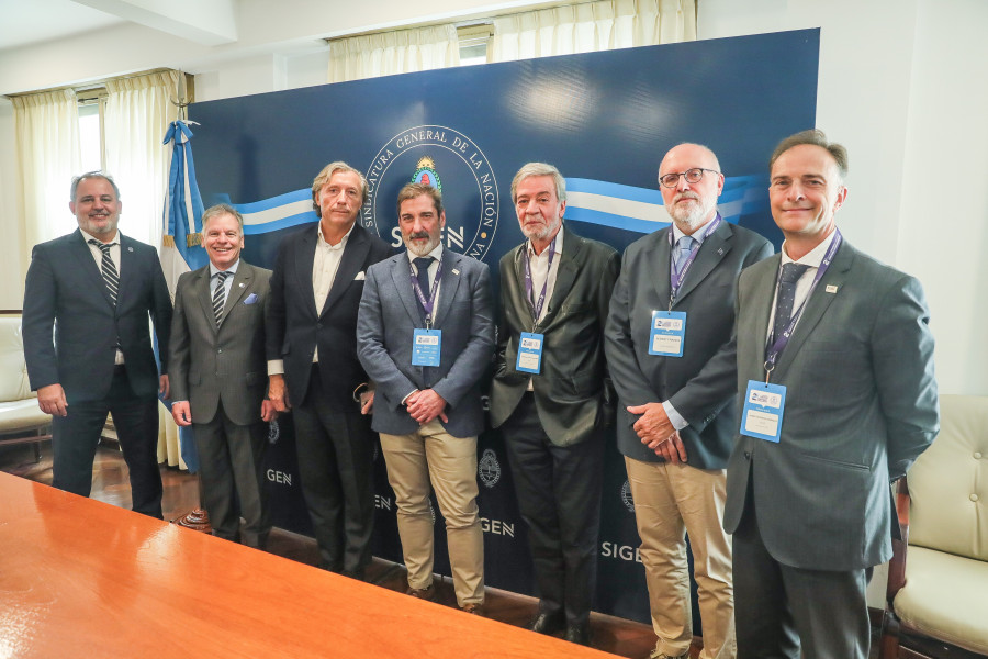 Sete indivíduos a posar sorridentes em frente a um cartaz, entre os quais se encontra o Inspetor-Geral