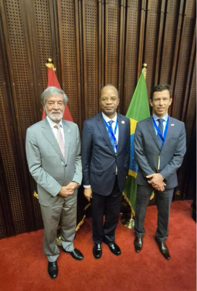 Três representantes das delegações portuguesa, angolana e brasileira posam com 2 bandeiras atrás de si