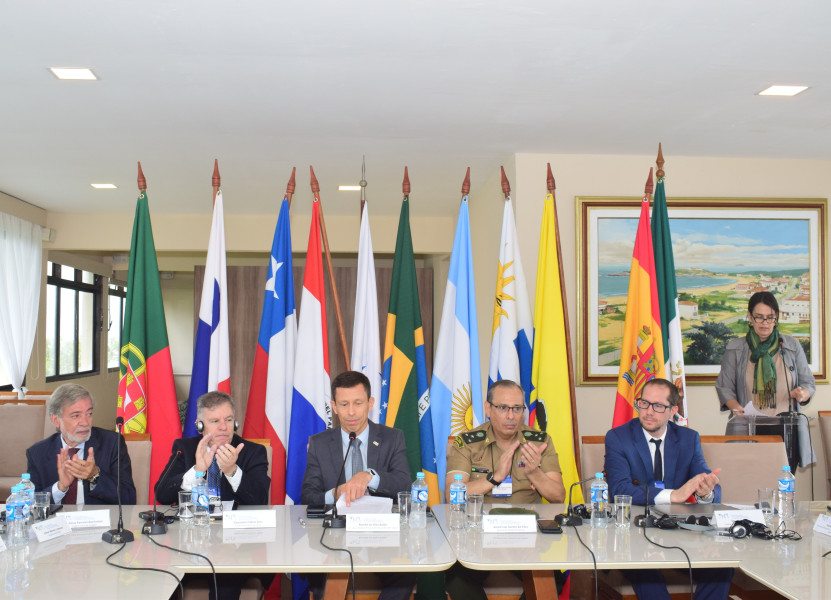 Inspetor-Geral com 4 pessoas sentados atrás de uma mesa de conferência à frente de várias bandeiras nacionais visíveis ao fundo