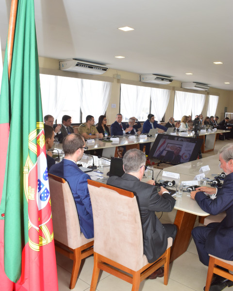 Vista da mesa de reunião numa sala com vários participantes sentados com a bandeira portuguesa em primeiro plano