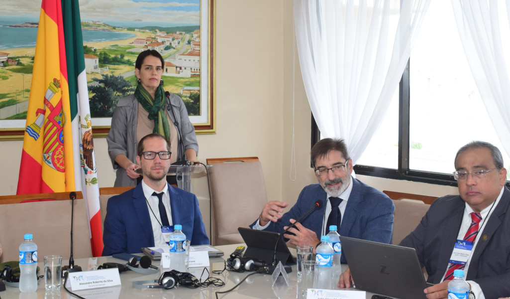 Três homens estão sentados e uma mulher está de pé atrás deles numa sala de reunião