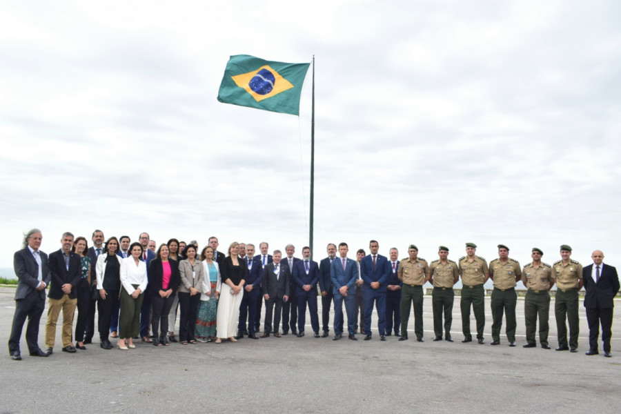Grupo de pessoas em pé no exterior para uma fotografia formal, com a bandeira do Brasil a voar ao vento num mastro alto ao centro.