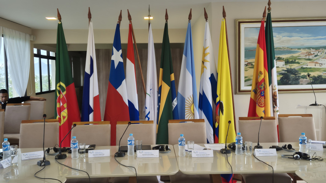 Mesas da reunião com garrafas de água e copos, com as bandeiras de vários países visíveis ao fundo