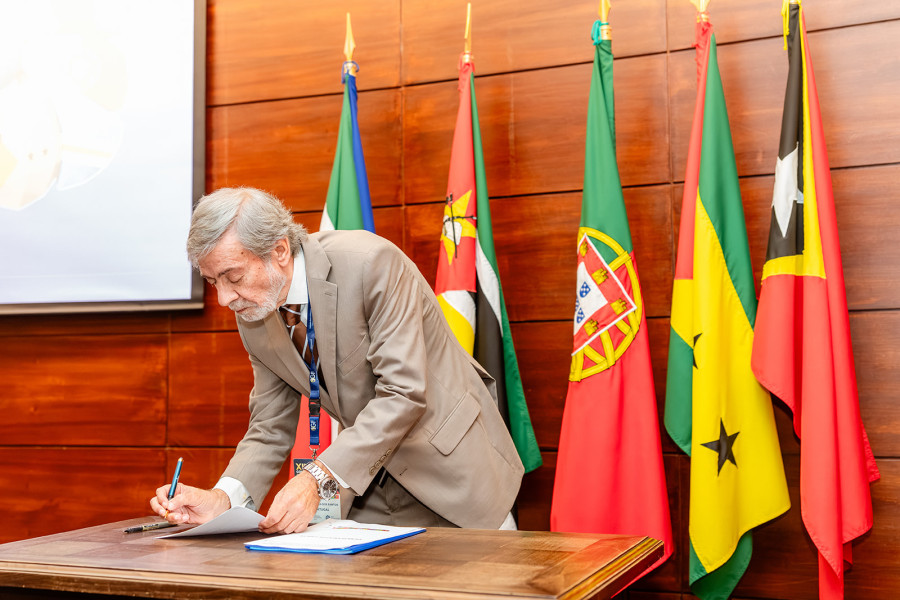 Assinatura por parte do IG de um documento que se encontra em cima de uma mesa em frente das bandeiras dos países membros da CPLP.