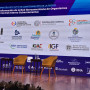 Palco do congresso onde se podem ver 2 cadeiras vazias e 2 mesas de apoio à frente de uma imagem gigante projetada do cartaz do congresso