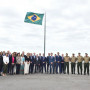 Grupo de pessoas em pé no exterior para uma fotografia formal, com a bandeira do Brasil a voar ao vento num mastro alto ao centro.