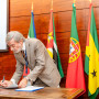 Assinatura por parte do IG de um documento que se encontra em cima de uma mesa em frente das bandeiras dos países membros da CPLP.