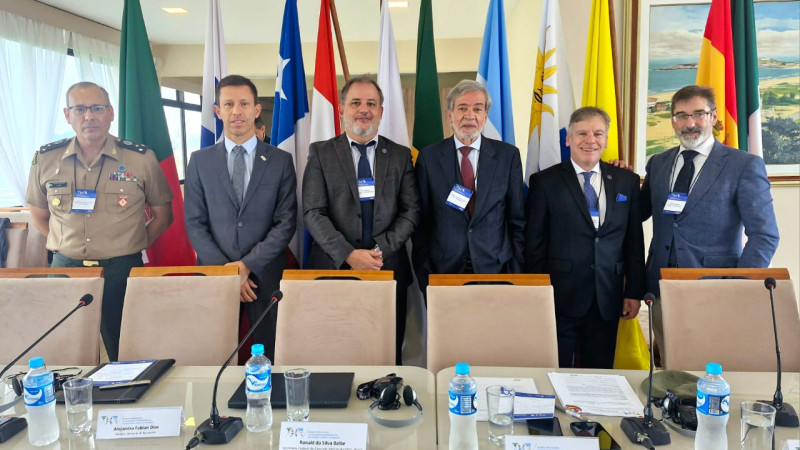 Inspetor-Geral com 5 pessoas atrás de uma mesa de conferência à frente de várias bandeiras nacionais visíveis ao fundo