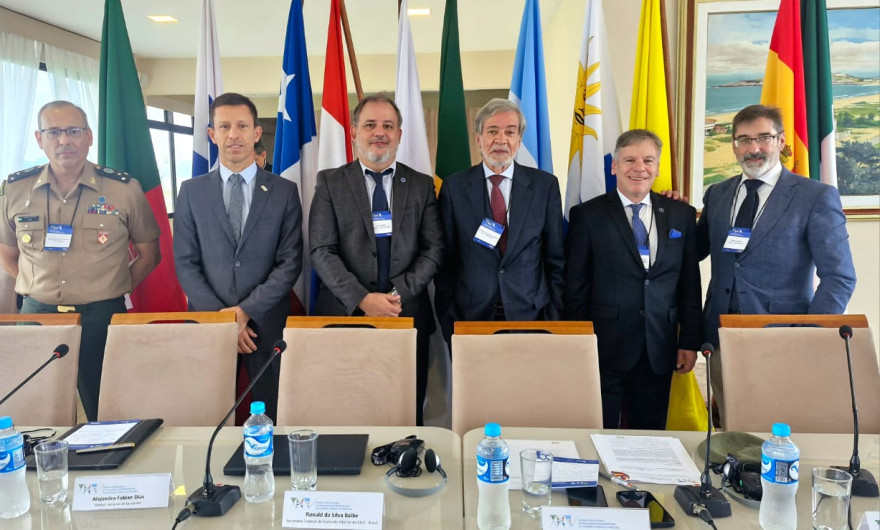 Inspetor-Geral com 5 pessoas atrás de uma mesa de conferência à frente de várias bandeiras nacionais visíveis ao fundo
